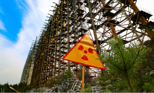 A triangular radiation warning sign stands near a massive rusting metal structure, resembling a radar array, with scattered debris and small trees in a blue sky setting.